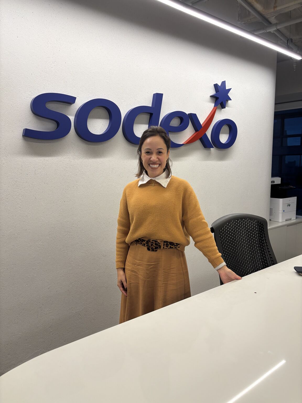 Colaboradora da Sodexo sorri em ambiente corporativo, posicionada em frente ao logotipo da empresa na parede, ao lado de uma mesa de escritório, representando acolhimento, profissionalismo e proximidade no ambiente de trabalho.
