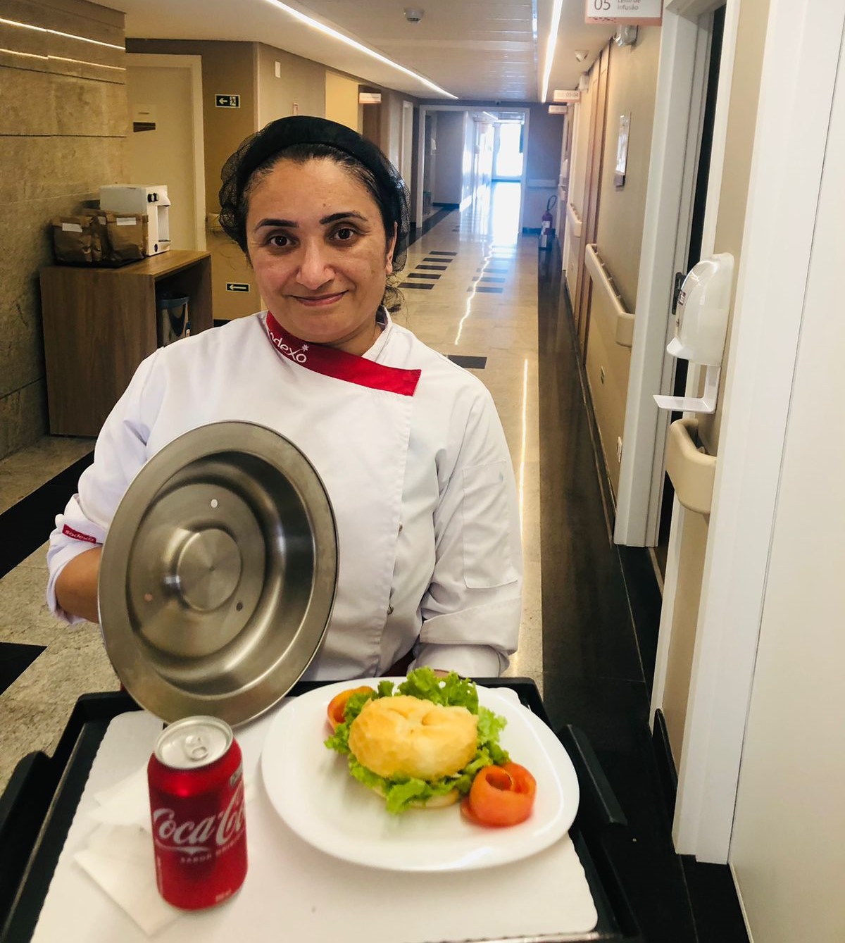 Chef Kátia vestindo um uniforme branco e vermelho da Sodexo, em um corredor iluminado de hospital, segurando uma bandeja com um prato contendo um sanduíche decorado com alface e tomate. 