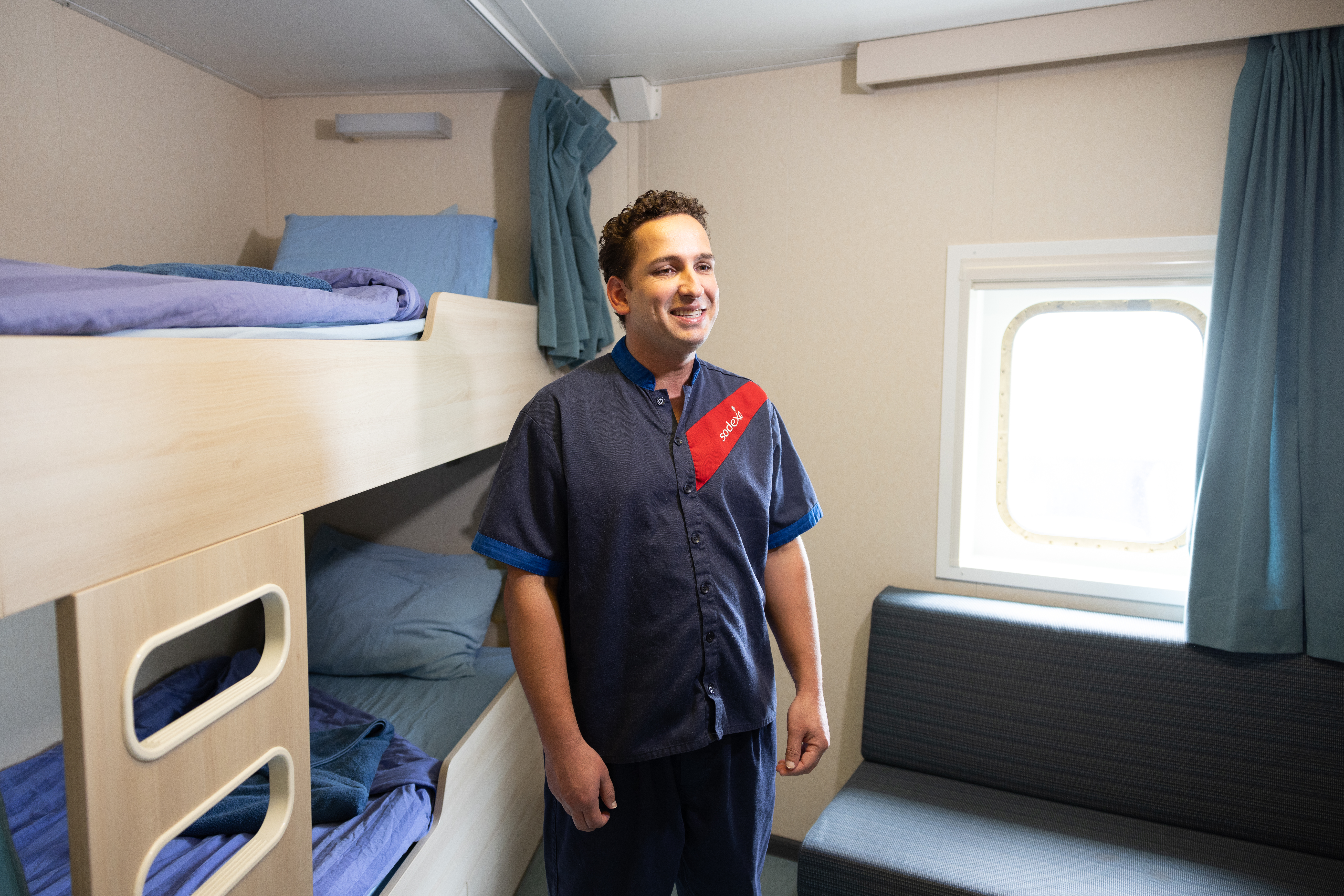 Profissional da Sodexo em uniforme azul sorri dentro de um camarote offshore, ao lado de beliche arrumado e janela com cortina azul. 