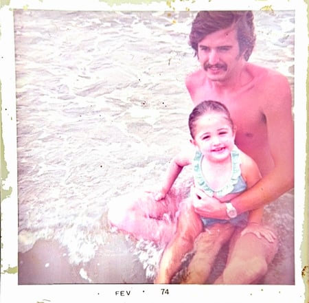 Homem e criança sorrindo e sentados na beira do mar. A imagem tem aparência antiga e está datada de fevereiro de 1974.