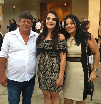 Três pessoas posam sorrindo para a foto em frente a um prédio iluminado, sugerindo um evento formal ou celebração. À esquerda, um homem de camisa branca e calça jeans; ao centro, uma mulher com vestido rendado em preto e bege; à direita, uma mulher com vestido bege e preto