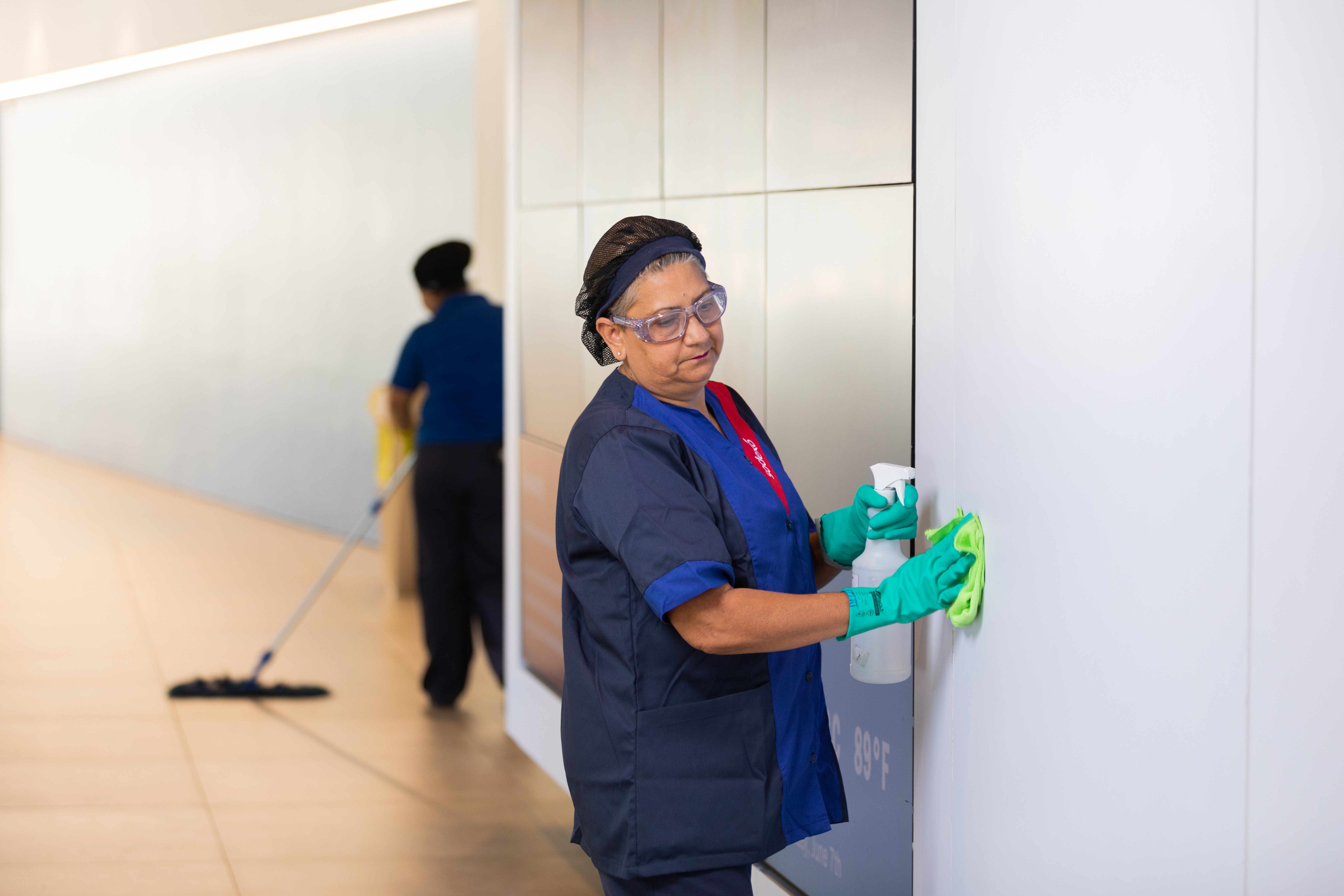 Funcionária da Sodexo realiza a limpeza de um ambiente, vestindo uniforme azul escuro com detalhes vermelhos, luvas verdes e óculos de proteção. Ela segura um borrifador e um pano branco. 
