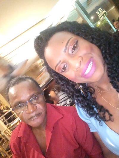 Selfie de duas pessoas sorrindo em um ambiente interno iluminado. À direita, está uma mulher com cabelos cacheados e batom rosa, enquanto à esquerda está um homem de óculos e camisa vermelha.