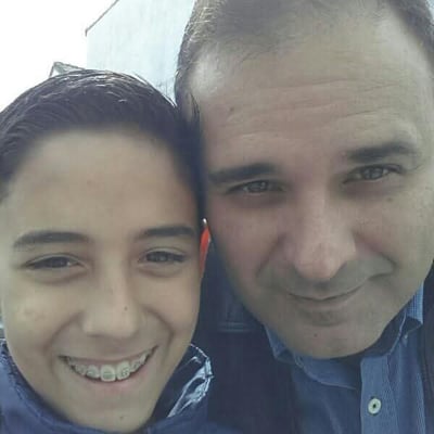 Selfie de um homem e um adolescente sorrindo lado a lado, em um ambiente externo durante o dia.