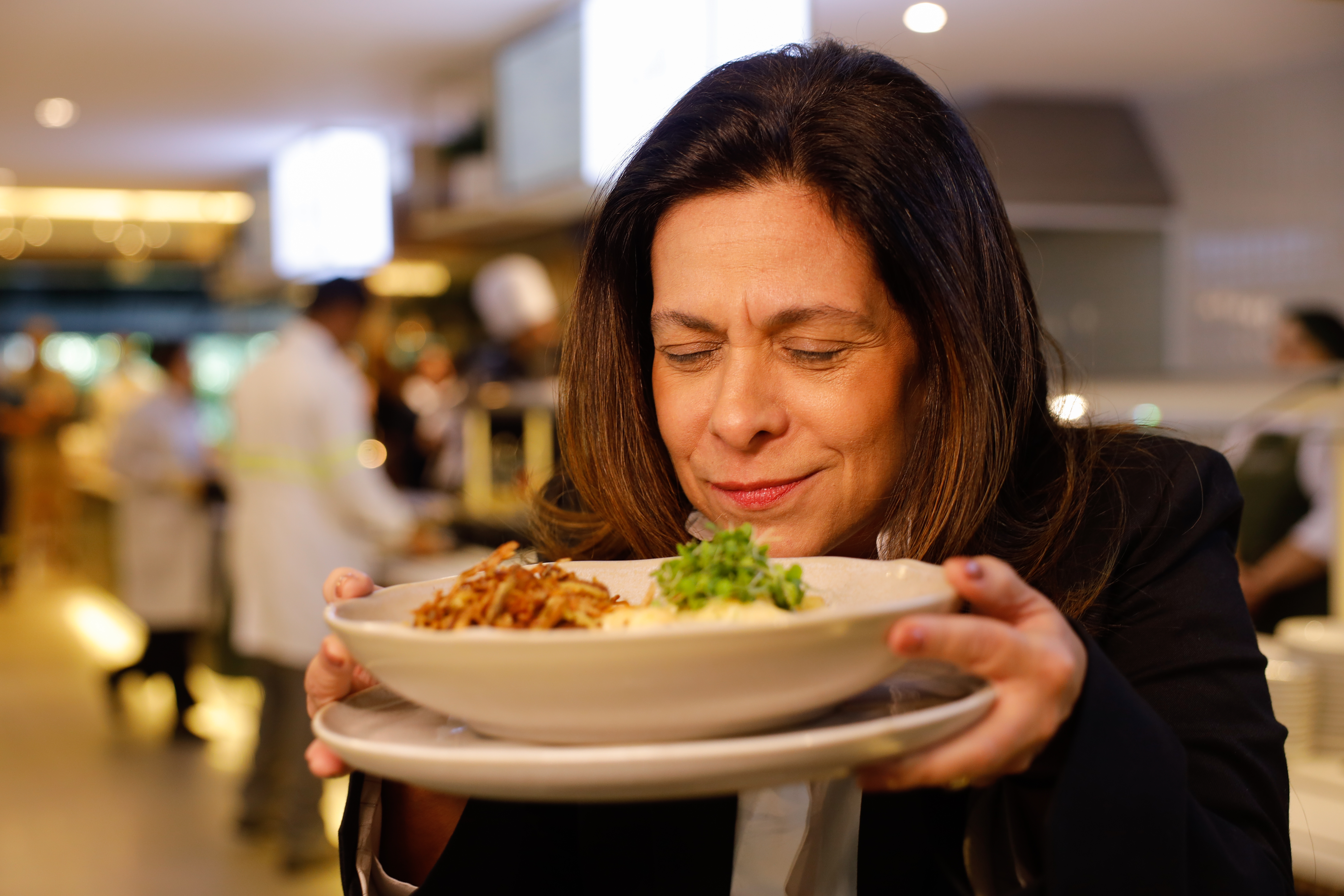 Mulher segura um prato próximo ao rosto e aprecia o aroma da refeição em um restaurante corporativo. 