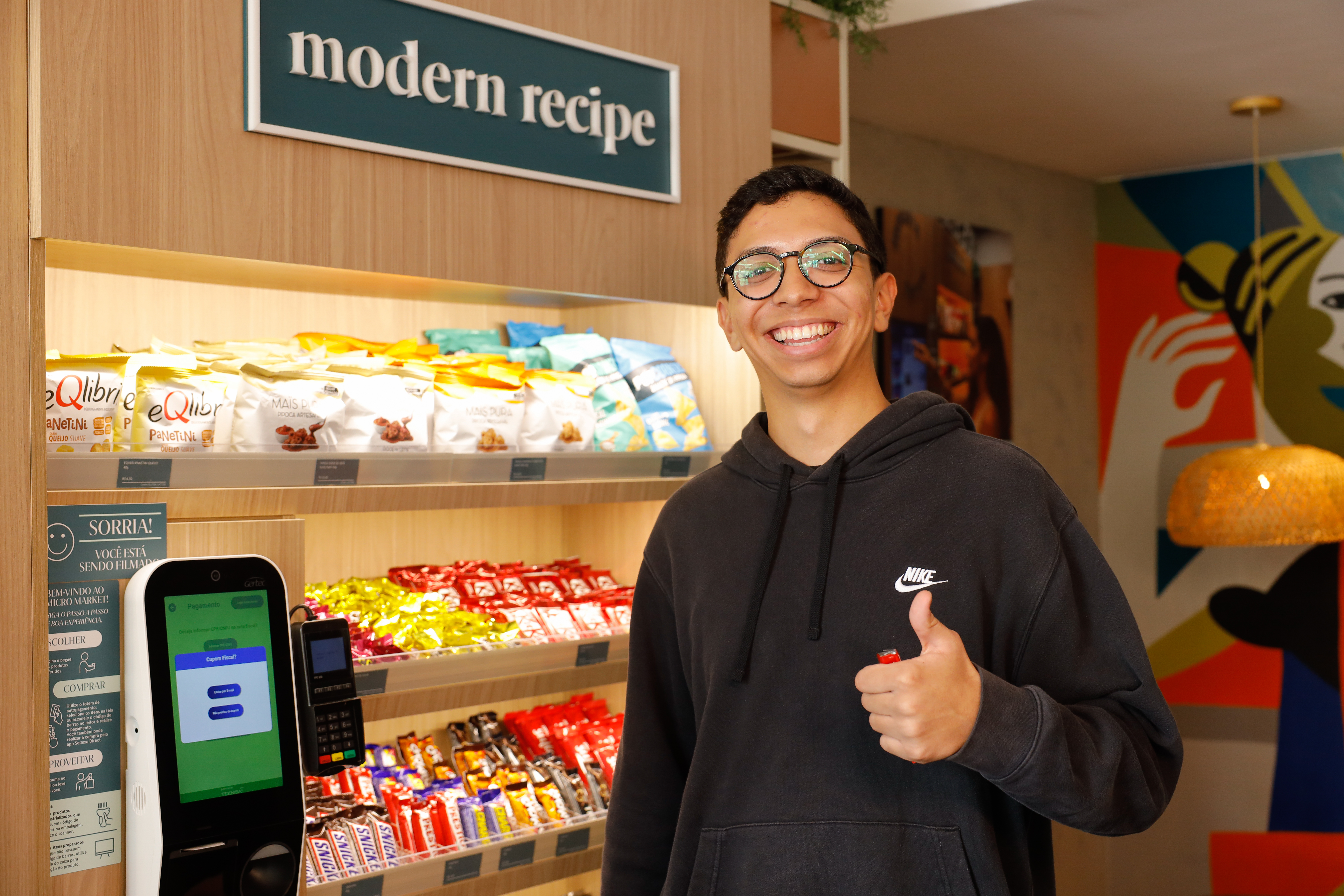 Homem sorri e faz sinal de positivo ao lado de um terminal de autoatendimento e de prateleiras com snacks em um micromercado Modern Recipe. 