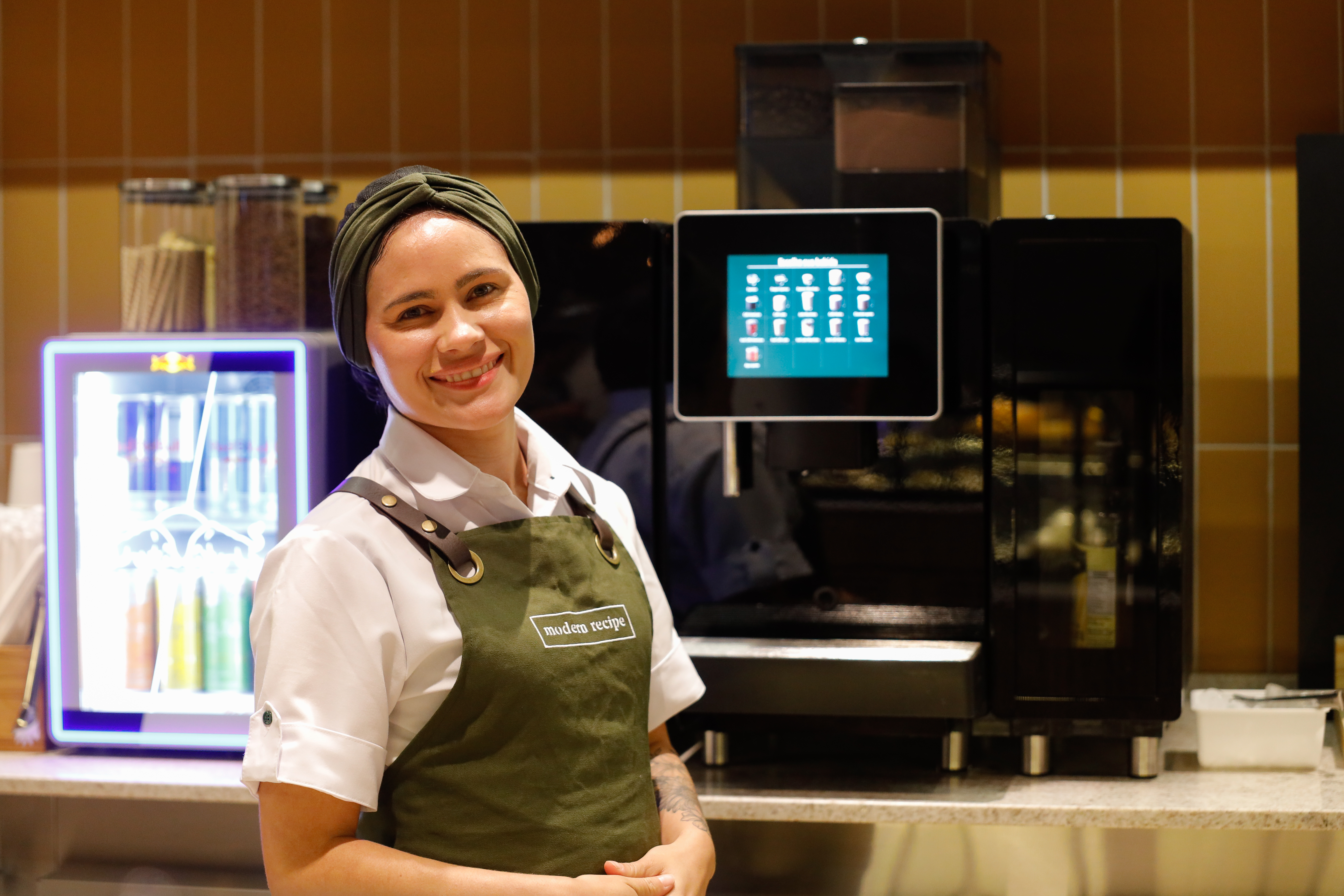 Colaboradora sorrindo em frente a máquinas de bebidas, usando uniforme e avental verde da Modern Recipe.