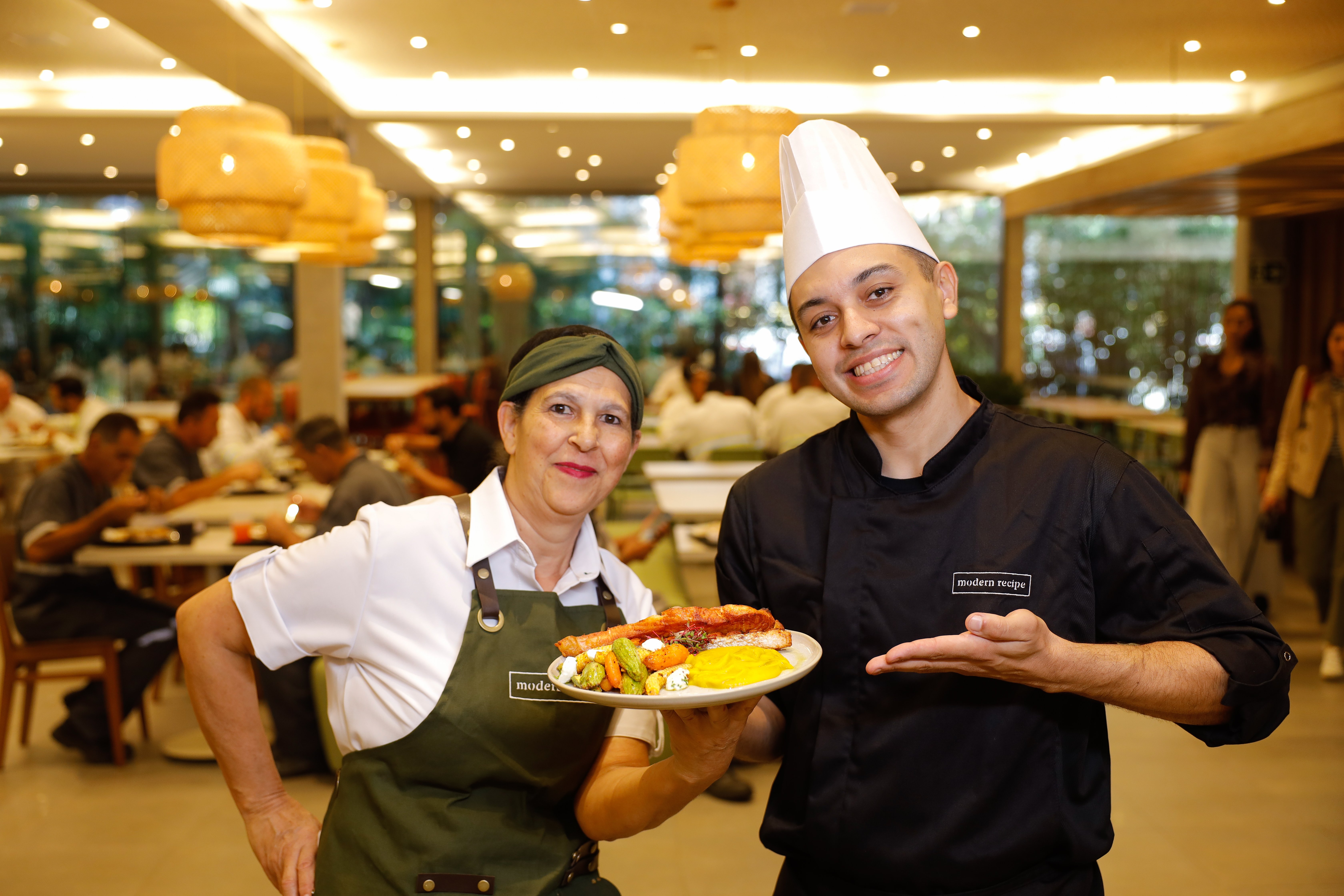 Dois colaboradores do Modern Recipe sorriem enquanto apresentam um prato de refeição no restaurante.