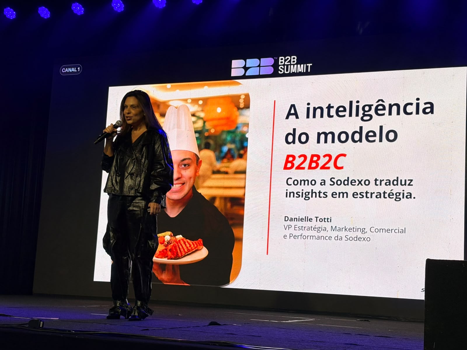 Palestrante no palco do B2B Summit 2026, em frente a um telão com o título “A inteligência do modelo B2B2C: como a Sodexo traduz insights em estratégia”. 