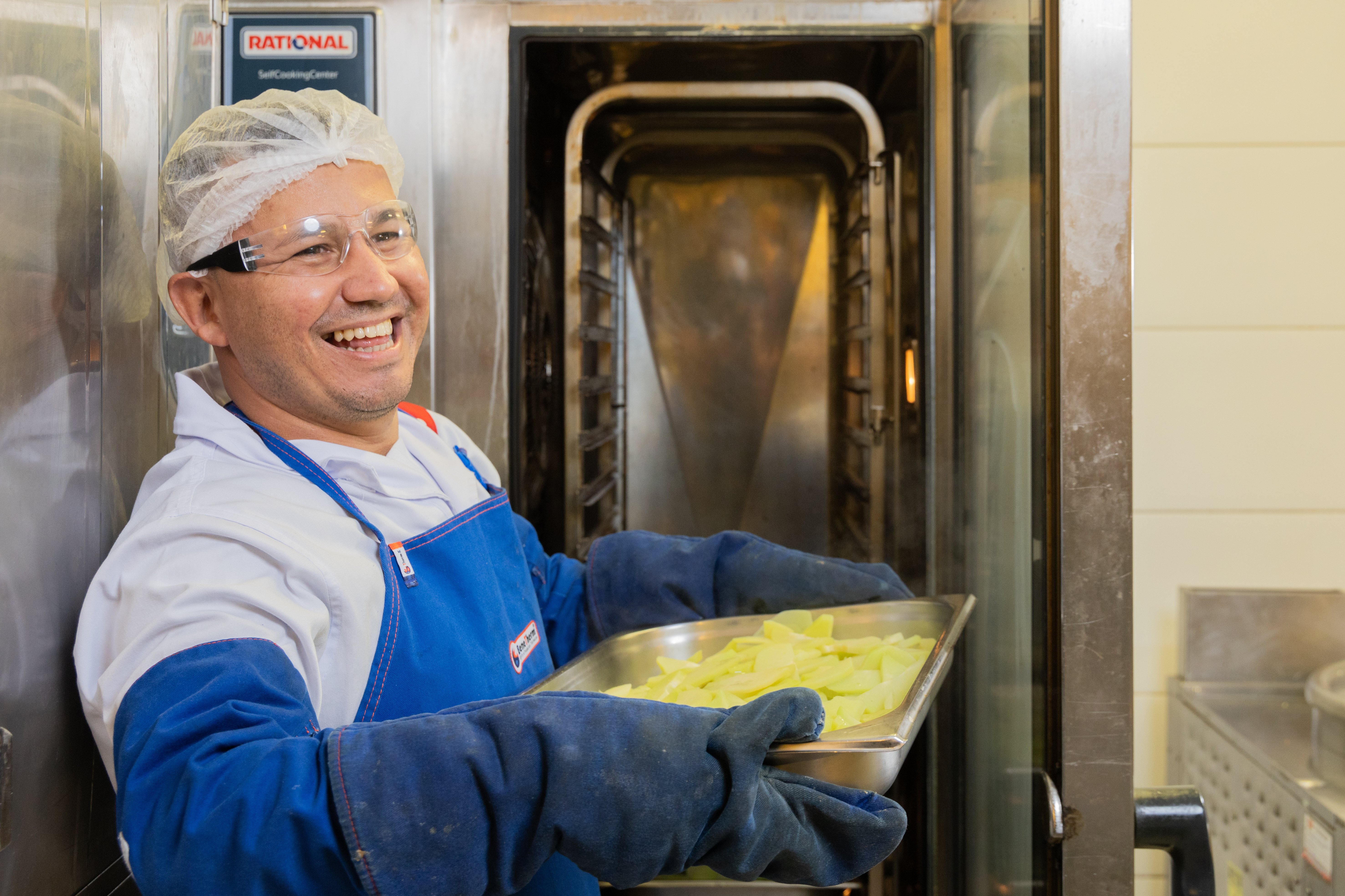 Colaborador sorrindo em uma cozinha profissional, usando equipamentos de proteção e segurando uma bandeja com batatas em frente a um forno industrial.