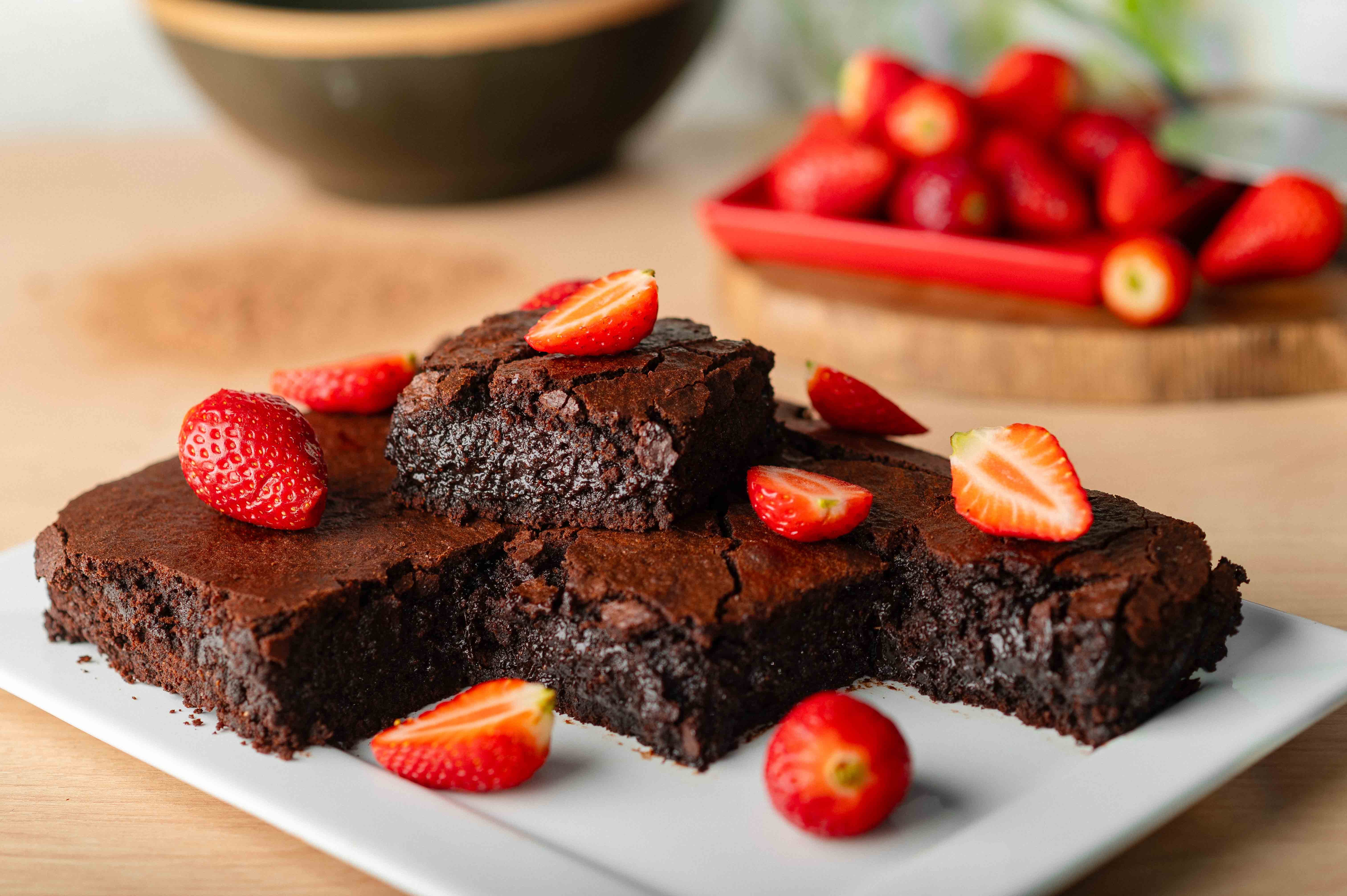 Brownies de chocolate cortados em quadrados e servidos em prato branco, decorados com morangos frescos. Ao fundo, há mais morangos sobre uma bandeja vermelha.