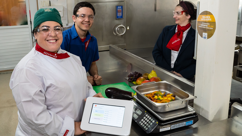 Três profissionais da Sodexo sorrindo em uma cozinha profissional, ao lado de uma balança com alimentos e um tablet utilizado para monitoramento.