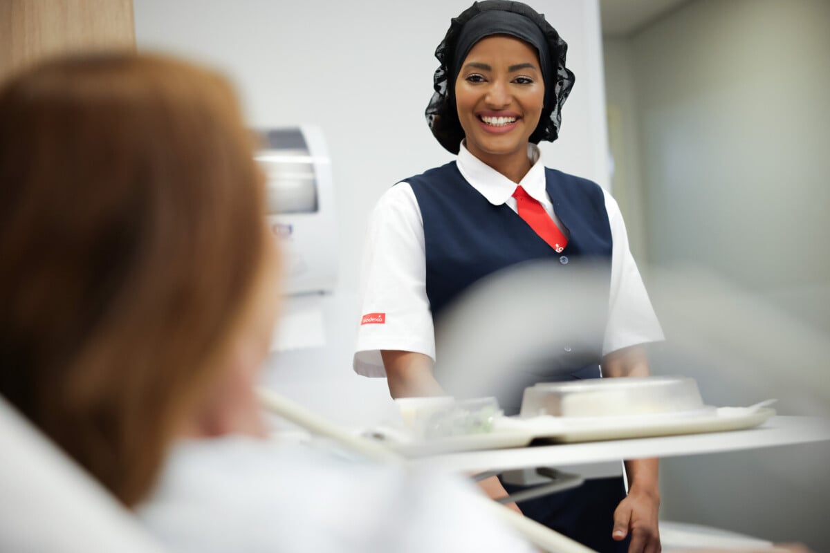 Profissional da Sodexo sorri enquanto entrega uma bandeja com refeição a uma paciente em um ambiente hospitalar, reforçando uma experiência de cuidado e acolhimento. 
