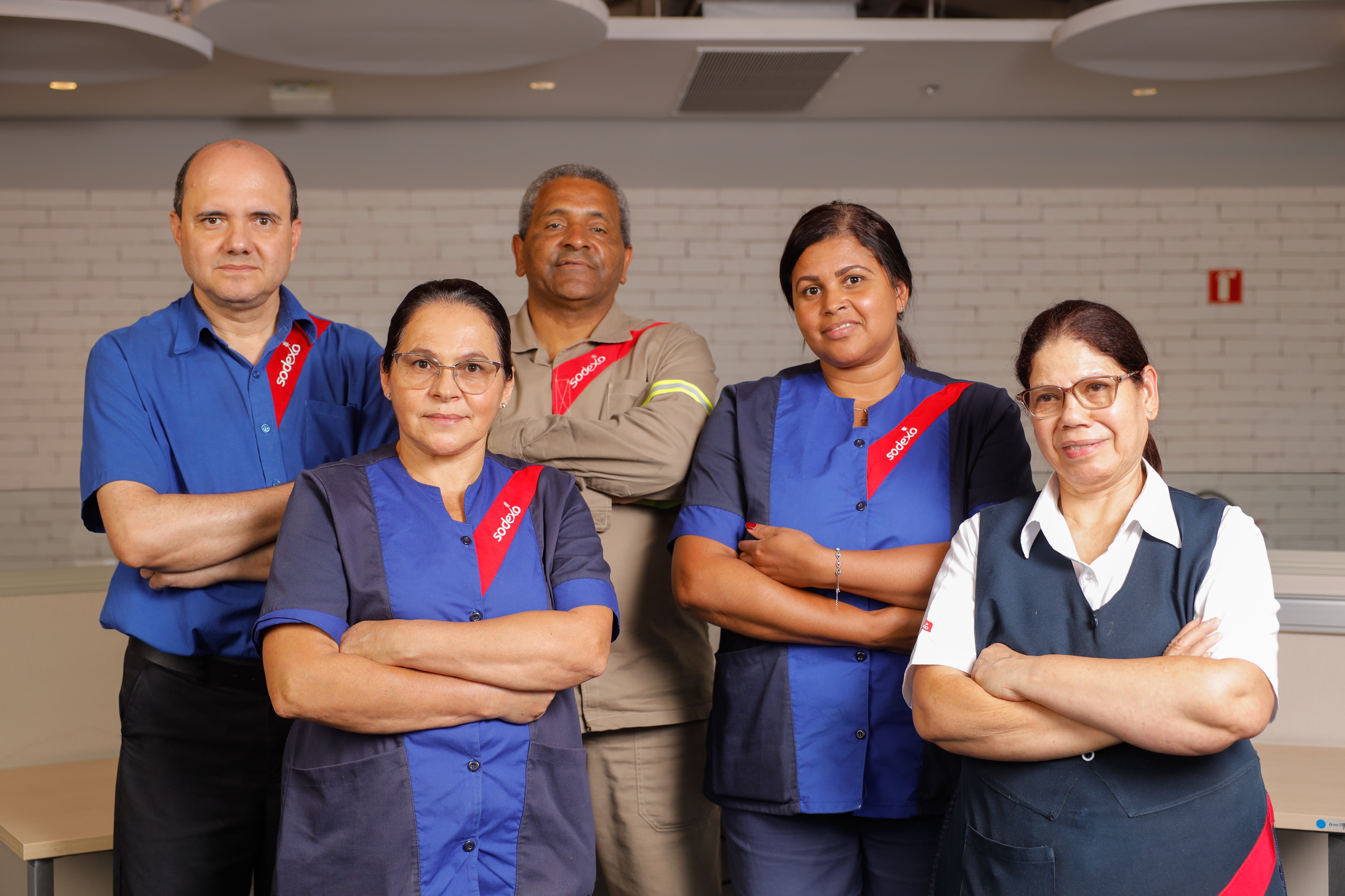 Cinco colaboradores da Sodexo posam para foto em um ambiente interno, vestindo uniformes da empresa em tons de azul, cinza e bege. Todos estão de braços cruzados, olhando para a câmera. 
