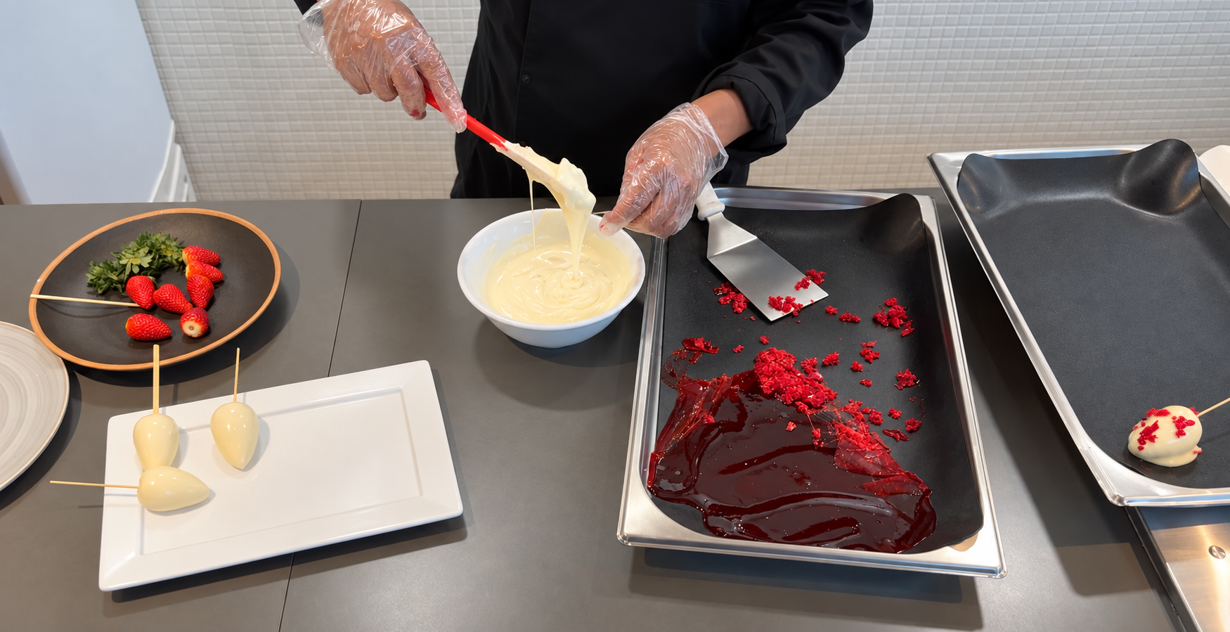 Pessoa prepara morangos cravejados, mergulhando os doces em chocolate branco ao lado de morangos frescos e pedaços de caramelo vermelho.