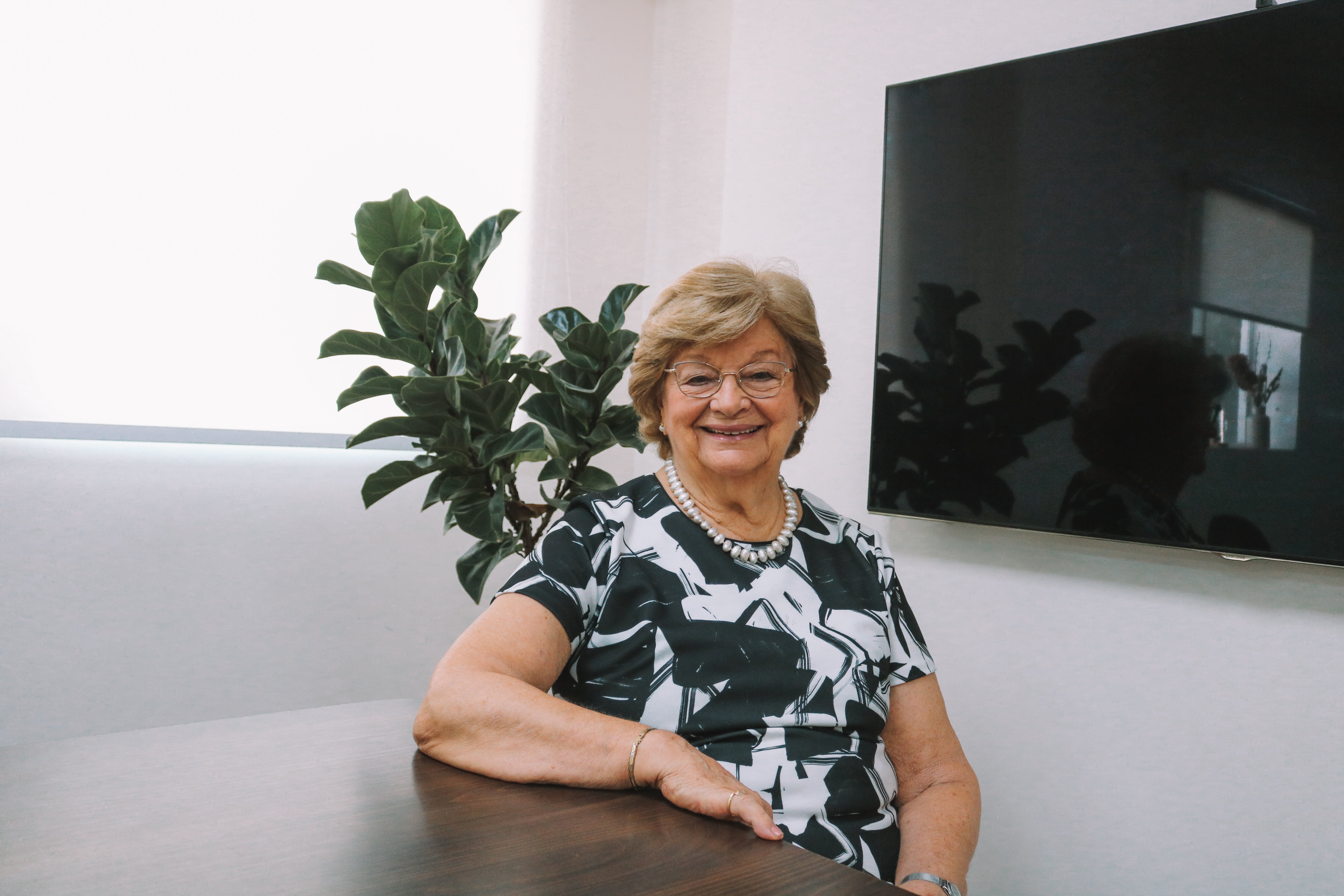 Mulher sorridente, de óculos e cabelo curto, sentada à mesa em um ambiente corporativo. Ao fundo, há plantas, uma janela com persiana e uma televisão fixada na parede. 