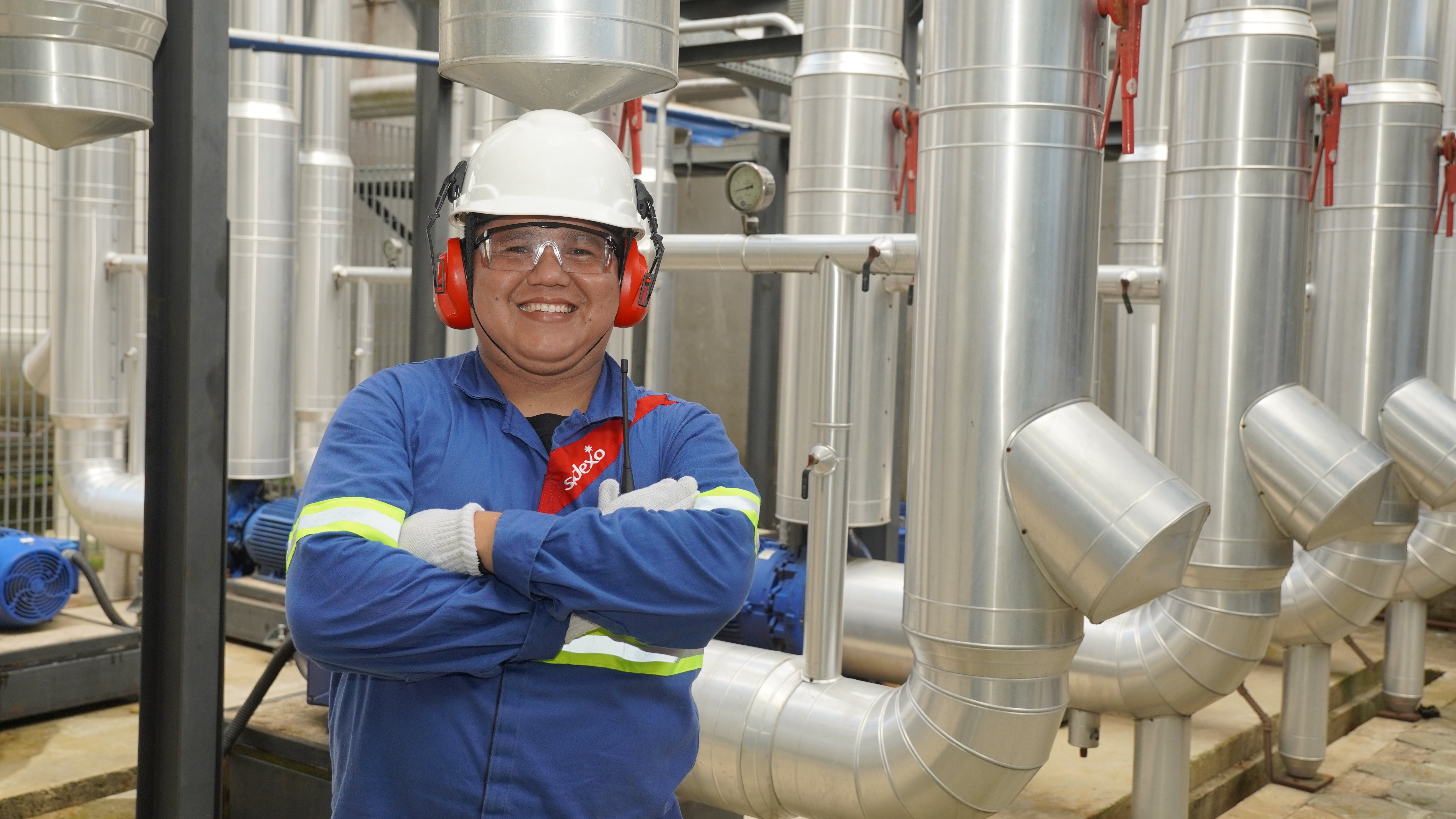 Profissional da Sodexo com uniforme azul e equipamentos de proteção individual posa em uma área industrial, cercada por tubulações e equipamentos.