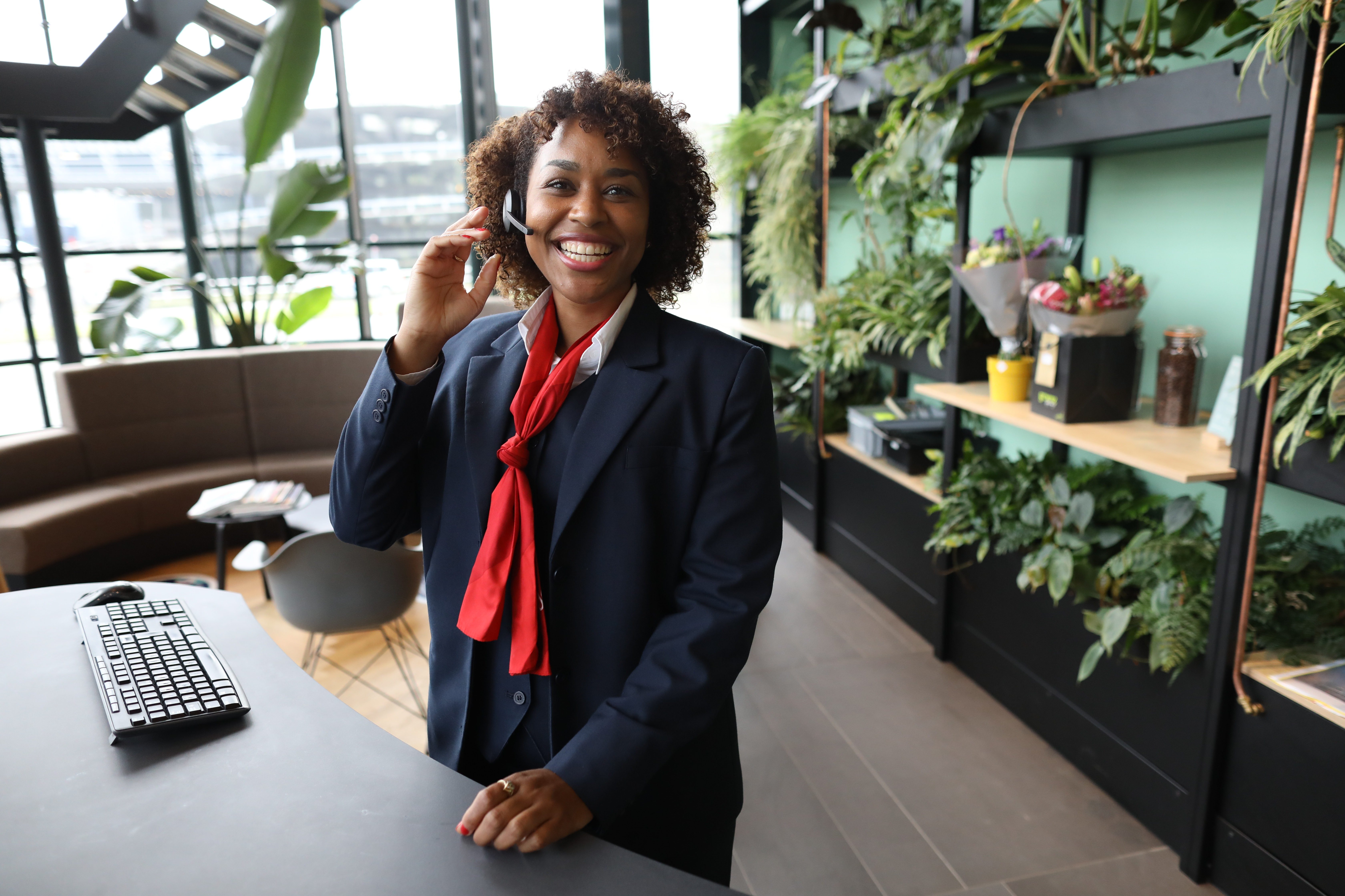 Mulher sorridente, vestindo uniforme social azul com lenço vermelho e fone de ouvido, atende no balcão de recepção em ambiente moderno com plantas e iluminação natural.