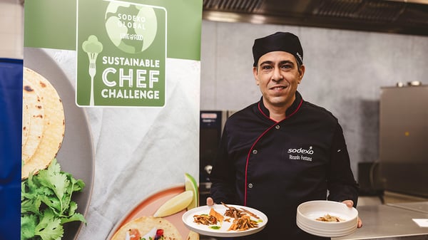 Chef sorridente com uniforme preto da Sodexo segura dois pratos com alimentos em frente a banner do “Sustainable Chef Challenge”, em cozinha industrial. 