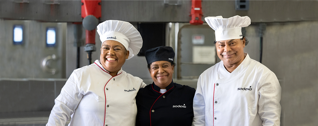 Três chefs da Sodexo uniformizados e sorrindo em uma cozinha industrial, transmitindo profissionalismo e espírito de equipe.