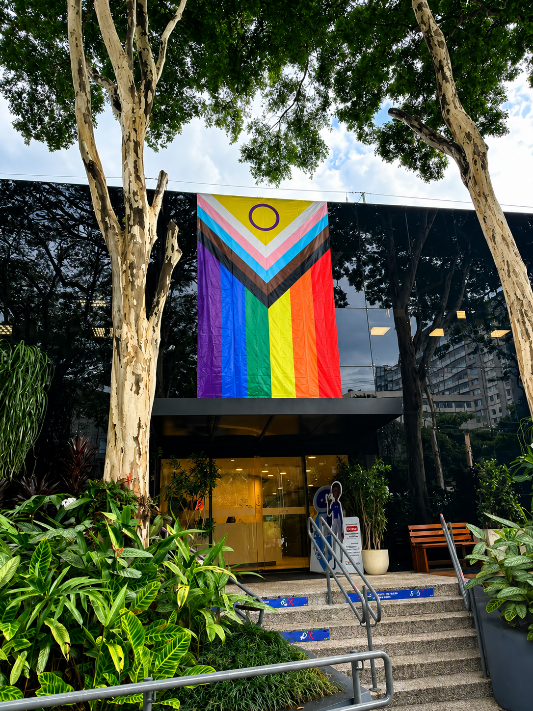 Fachada da sede da Sodexo Brasil, com uma grande bandeira LGBTI+ hasteada em destaque em comemoração ao Mês do Orgulho. A entrada do prédio aparece cercada por árvores e plantas, reforçando o compromisso da empresa com diversidade, inclusão, respeito e pertencimento.