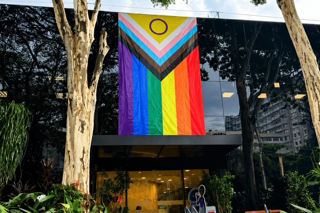 Fachada da sede da Sodexo Brasil, com uma grande bandeira LGBTI+ hasteada em destaque em comemoração ao Mês do Orgulho. A entrada do prédio aparece cercada por árvores e plantas, reforçando o compromisso da empresa com diversidade, inclusão, respeito e pertencimento.