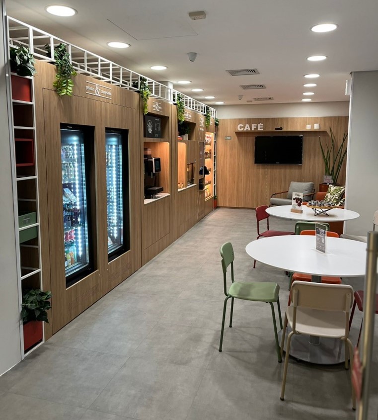 Espaço interno de um micromercado da Sodexo, com design moderno e acolhedor, apresentando prateleiras com produtos variados, refrigeradores, máquinas de café e mesas com cadeiras para momentos de pausa e integração.