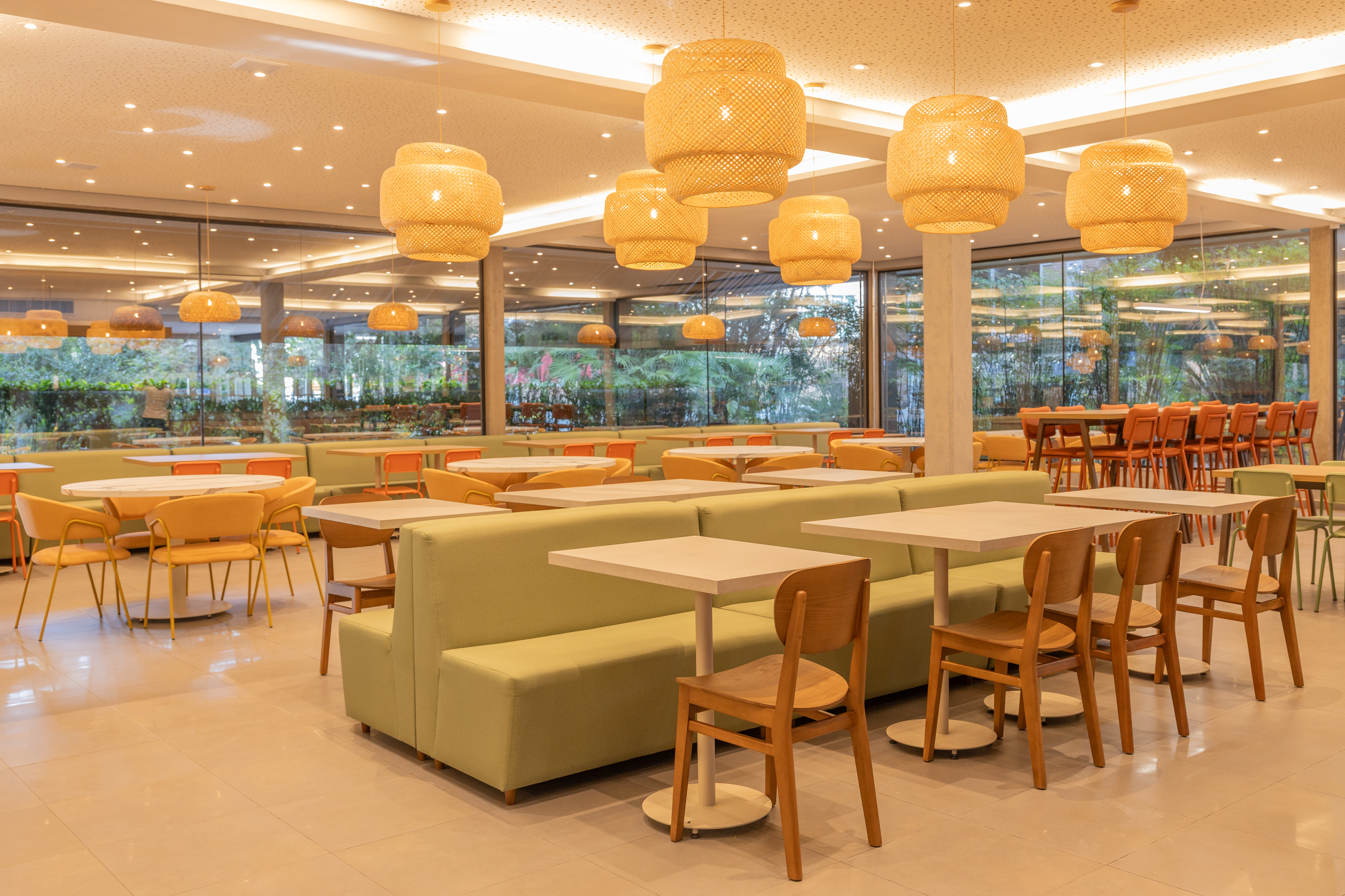 Restaurante corporativo amplo e iluminado, com mesas, cadeiras de madeira, sofás verdes, luminárias pendentes em tons naturais e grandes janelas de vidro com vista para área externa arborizada.