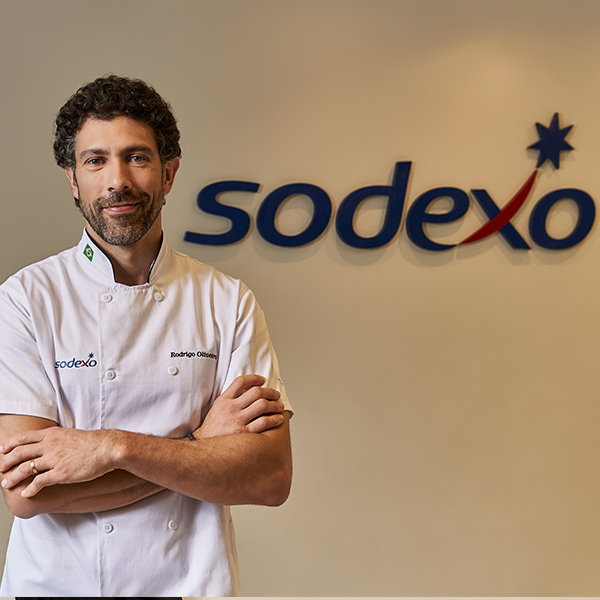 Chef Rodrigo Oliveira com os braços cruzados ao lado do logo da Sodexo na parede.