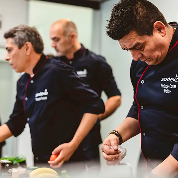 Três chefs em uniformes da Sodexo estão trabalhando em uma cozinha. Um está segurando um recipiente, outro parece estar cortando algum alimento, e o terceiro está focado em uma tarefa. O fundo está desfocado.