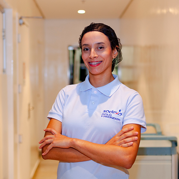 Uma jovem funcionária da Sodexo, com touca e avental azul marinho, sorri enquanto serve comida em um buffet. O buffet possui diversos recipientes com opções de salada, como tomate, milho e outros vegetais coloridos. A cena se passa em um refeitório ou restaurante, com mesas e outros clientes ao fundo, levemente desfocados.