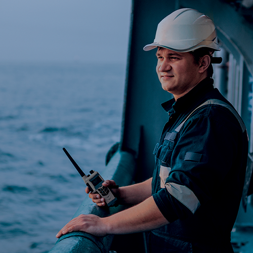 Trabalhador offshore usando capacete branco e uniforme de segurança, segurando um rádio comunicador enquanto observa o mar a partir da embarcação.