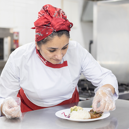 Chef Kátia finaliza a decoração de um prato. Ela sorri e usa um uniforme branco e vermelho da Sodexo.  