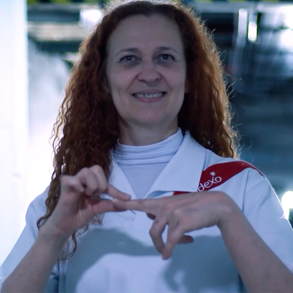 Mulher ruiva, vestindo uniforme branco da Sodexo, sorri enquanto faz um sinal com as mãos em Língua Brasileira de Sinais (Libras). Ela está em um ambiente corporativo.