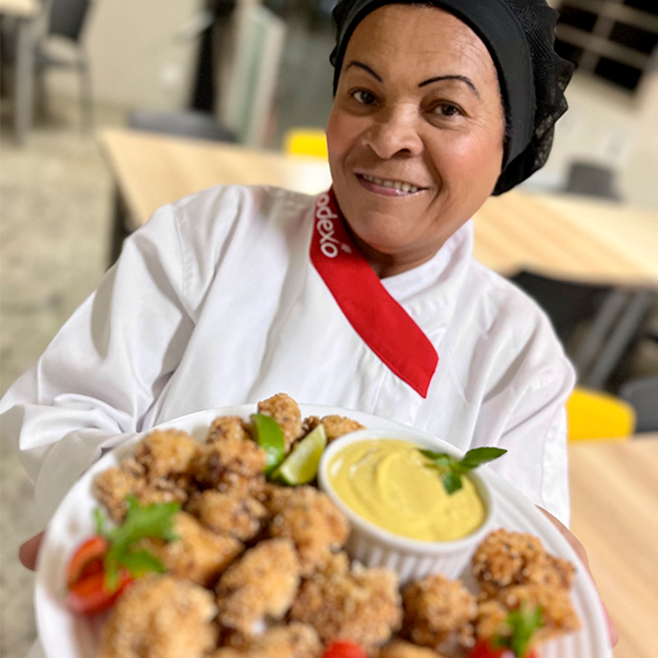 Cozinheira da Sodexo, Maria Nilza, segurando um prato de pedaçoes crocantes de couve-flor com maionese de abacate, decorado com fatias de limão e tomates cereja