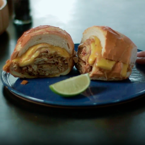 Sanduíche de pão francês recheado com pernil desfiado e queijo derretido, servido em um prato azul com uma fatia de limão ao lado.