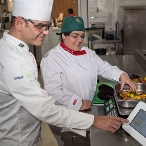Um homem e uma mulher estão juntos na cozinha, envolvidos em atividades culinárias, rodeados por utensílios de cozinha.