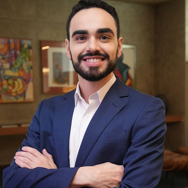 Um advogado com terno azul, camisa social branca, com os braços cruzados e sorrindo.