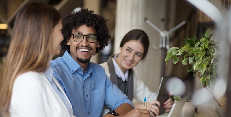 Three coworkers at a desk, one speaking while others smile, with laptop, notebook, and plant in office setting