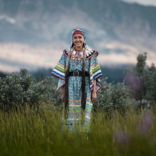 A woman in traditional attire stands gracefully in a vibrant green field under a clear blue sky.