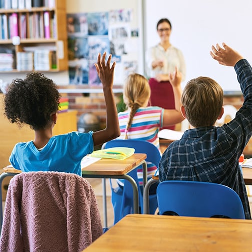 Copilot said: Élèves assis à leur bureau dans une salle de classe, levant la main tandis qu’un enseignant anime une leçon interactive dans un environnement d’apprentissage lumineux et bien organisé.