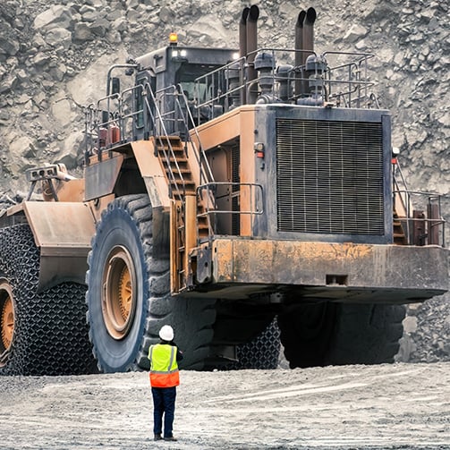 Large haul truck in an open-pit mine towers over a worker in high-visibility safety gear.