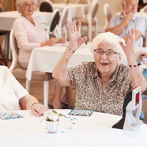 Older adults play bingo at tables in a community dining room, with one participant raising both hands.