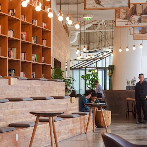 Modern open workspace with wooden seating steps, hanging lights, bookshelves, and people working and talking near a café-style counter.