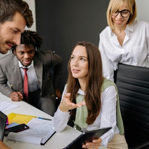 A group of people gathered around a table in an office, reviewing documents and discussing work while using a tablet and laptops.