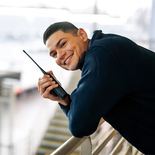 Person leaning on a railing while holding a two way radio in an outdoor setting with steps and structures in the background.