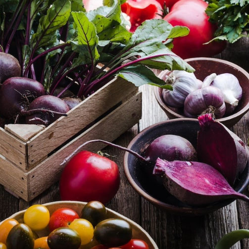 Assortment of fresh vegetables including beets, tomatoes, garlic, leafy greens, and colorful cherry tomatoes arranged on a rustic wooden table, highlighting healthy ingredients and sustainable food sourcing.
