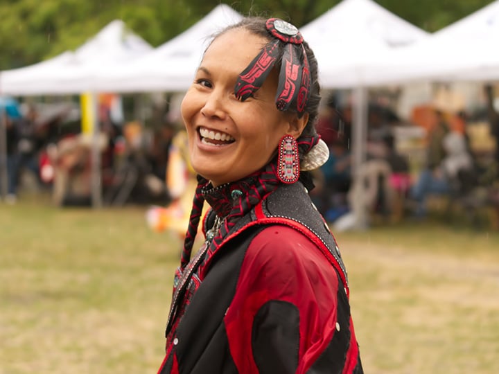 An indigenous woman in traditional attire smiles toward the camera at an outdoor gathering with white tents and people in the background.