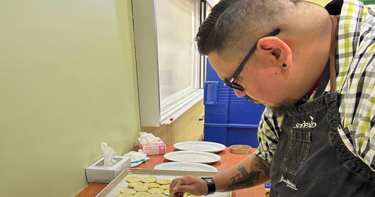Chef wearing an apron and glasses placing sliced food onto a tray in a kitchen workspace, with plates, utensils, and containers arranged on a counter nearby.