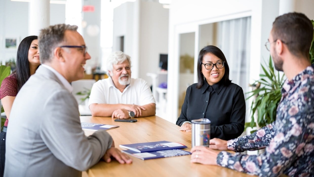 Group of people seated around a wooden table in a bright office space, discussing documents and collaborating during a meeting.
